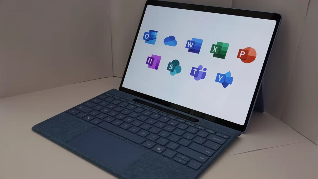 A Microsoft Surface with keyboard, its screen showing Microsoft 365 app icons including Outlook and OneDrive