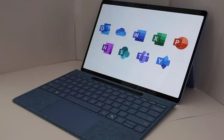 A Microsoft Surface with keyboard, its screen showing Microsoft 365 app icons including Outlook and OneDrive